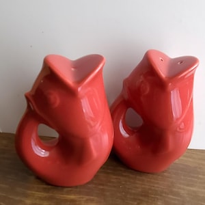 May include: Two red ceramic salt and pepper shakers. Each shaker is shaped like a fish with a heart-shaped opening at the top and a circular opening on the side. The shakers are glossy and sit on a wooden surface.