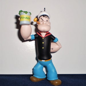 Może przedstawiać: Plastikowa figurka Popeye'a, marynarza, trzymającego puszkę szpinaku. Ma na sobie niebieską marynarską koszulę, czerwoną chustkę i biały marynarski kapelusz. Mruga okiem i trzyma fajkę w ustach.