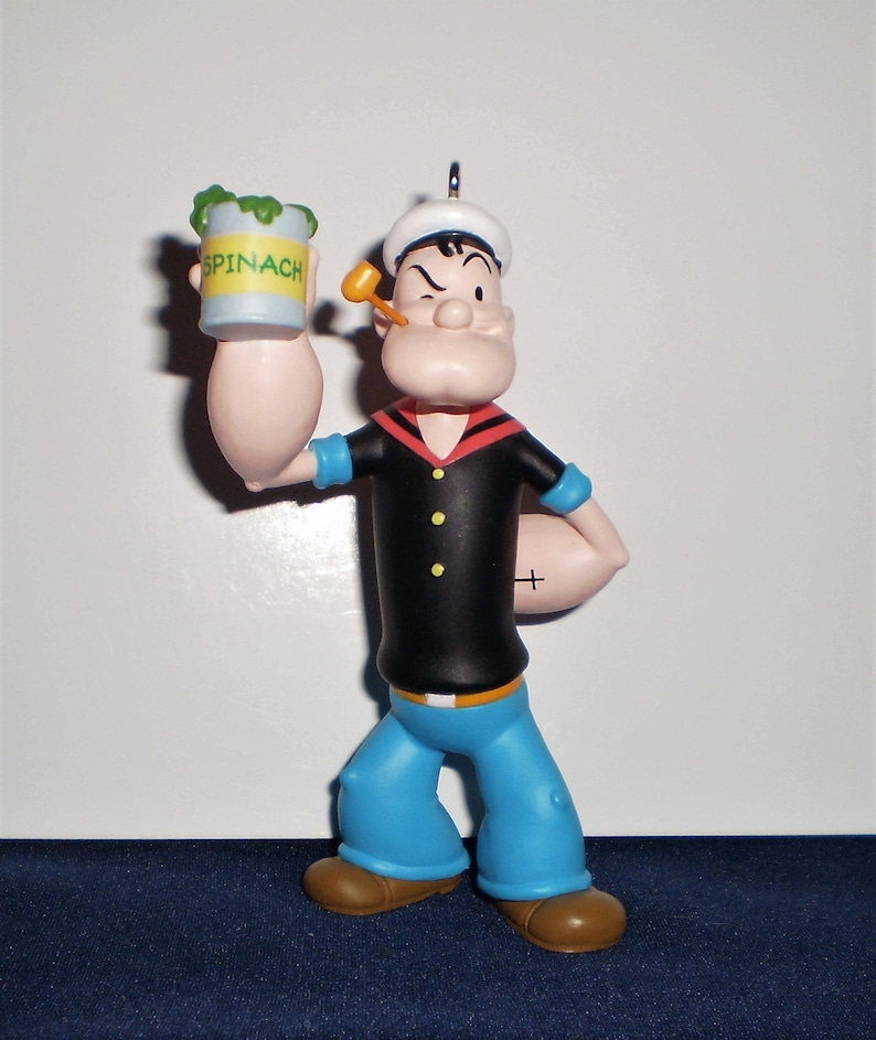 Może przedstawiać: Plastikowa figurka Popeye'a, marynarza, trzymającego puszkę szpinaku. Ma na sobie niebieską marynarską koszulę, czerwoną chustkę i biały marynarski kapelusz. Mruga okiem i trzyma fajkę w ustach.