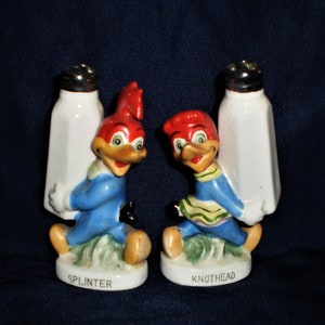 May include: A pair of ceramic salt and pepper shakers featuring the cartoon characters, Woody Woodpecker and his sidekick, Knothead. The shakers are painted in bright colors, with Woody wearing a blue jumpsuit and Knothead wearing a blue and green dress. The shakers are holding salt and pepper shakers in their hands. The base of each shaker is labeled with the character's name, "Splinter" and "Knothead".