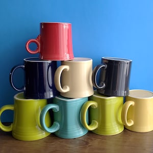 May include: A stack of ten ceramic mugs in various colors, including red, navy blue, beige, black, green, teal, and yellow. Each mug has a rounded handle and a simple, classic design. The mugs are arranged against a solid blue background.