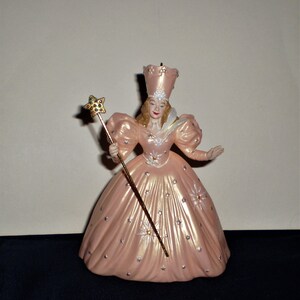 Peut inclure: Une figurine en porcelaine de la M&eacute;chante Sorci&egrave;re de l'Ouest du Magicien d'Oz. Elle porte une robe rose avec des &eacute;toiles argent&eacute;es et un grand chapeau pointu. Elle tient une baguette dor&eacute;e surmont&eacute;e d'une &eacute;toile.