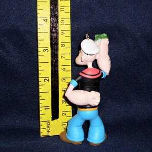Może przedstawiać: Plastikowa figurka Popeye'a, marynarza, trzymającego puszkę szpinaku w prawej ręce. Ma na sobie charakterystyczne niebieskie spodnie marynarskie, czarną koszulę z czerwonym kołnierzykiem i biały kapelusz marynarski. Figurka ma około 10 cm wysokości.