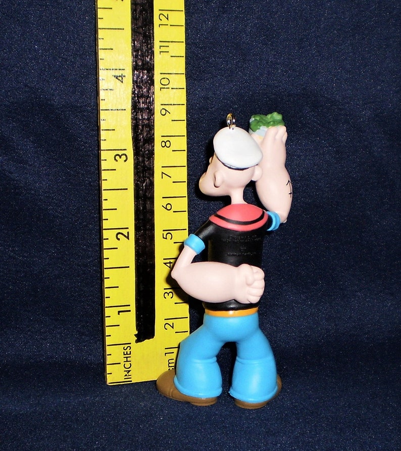 Może przedstawiać: Plastikowa figurka Popeye'a, marynarza, trzymającego puszkę szpinaku w prawej ręce. Ma na sobie charakterystyczne niebieskie spodnie marynarskie, czarną koszulę z czerwonym kołnierzykiem i biały kapelusz marynarski. Figurka ma około 10 cm wysokości.