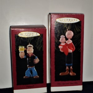 Może przedstawiać: Dwa czerwone pudełka z pozłacanymi obw&oacute;dkami zawierające świąteczne ozdoby z Popeyem i Oliwką. Na pudełkach widnieje napis "Keepsake Ornament".