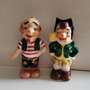 May include: A pair of vintage ceramic figurines, a man and a woman, with exaggerated features and colorful clothing. The man is wearing a red and white striped shirt, a green vest, blue pants, and yellow shoes. The woman is wearing a green jacket, a plaid skirt, pink pants, and brown shoes.