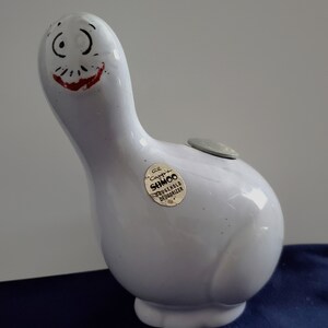 May include: White ceramic goose shaped piggy bank with a black and red painted face. The goose has a coin slot on its back and a sticker that reads "Ce Cappri SHMCO Household Deodorizer".