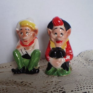 May include: Two vintage ceramic figurines of seated elves. One elf wears a white hat, red scarf, white shirt, green pants, and black shoes. The other elf has a red cap, yellow vest, red jacket, black pants, and holds a black pot.