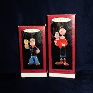 Może przedstawiać: Dwa czerwone pudełka z białymi obw&oacute;dkami zawierające świąteczne ozdoby w stylu vintage. Ozdoby przedstawiają Popeye'a, marynarza. Jedna ozdoba przedstawia Popeye'a trzymającego szklankę szpinaku. Druga ozdoba przedstawia Popeye'a trzymającego Oliwię. Pudełka są oznaczone "Keepsake Ornaments".