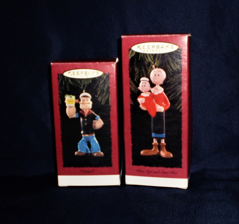 Może przedstawiać: Dwa czerwone pudełka z białymi obw&oacute;dkami zawierające świąteczne ozdoby w stylu vintage. Ozdoby przedstawiają Popeye'a, marynarza. Jedna ozdoba przedstawia Popeye'a trzymającego szklankę szpinaku. Druga ozdoba przedstawia Popeye'a trzymającego Oliwię. Pudełka są oznaczone "Keepsake Ornaments".