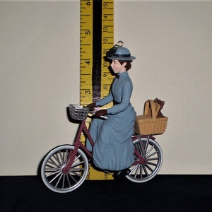 Peut inclure: Une figurine bleue et blanche d'une femme &agrave; v&eacute;lo avec un panier &agrave; l'arri&egrave;re. La femme porte une robe bleue et un chapeau. Le v&eacute;lo est rouge avec des pneus blancs.
