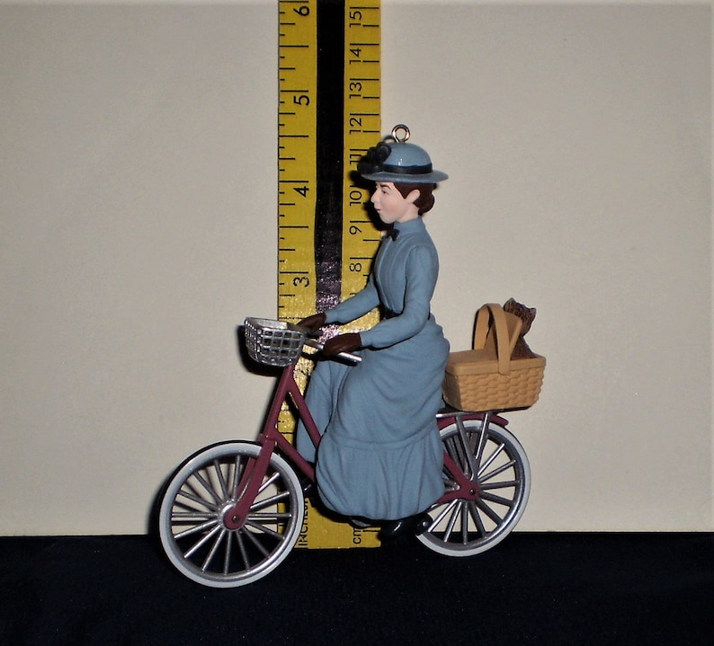 Peut inclure: Une figurine bleue et blanche d'une femme &agrave; v&eacute;lo avec un panier &agrave; l'arri&egrave;re. La femme porte une robe bleue et un chapeau. Le v&eacute;lo est rouge avec des pneus blancs.