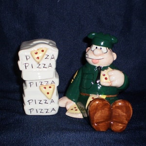 May include: A ceramic figurine of a cartoon pizza delivery person wearing a green uniform and holding a slice of pizza. The figurine is sitting down with its legs crossed. There are three stacked pizza boxes behind the figurine, each with the word "PIZZA" written on it.