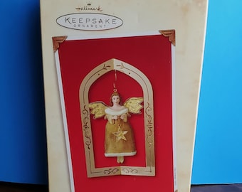 Peut inclure: Boîte d'ornement Hallmark Keepsake avec un ornement d'ange doré. L'ange a des ailes dorées et une étoile sur sa robe, sur fond rouge. La boîte indique "Glad Tidings" et est dans une boîte de couleur crème.