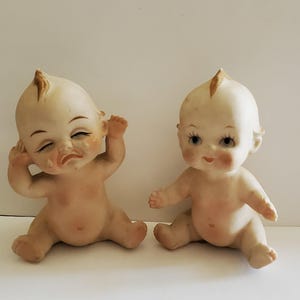 May include: Two porcelain baby dolls, one with a sad expression and the other with a happy expression. Both dolls are sitting with their legs crossed and have painted eyes and hair.