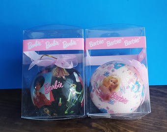 Peut inclure: Deux boules de Noël sur le thème de Barbie dans des boîtes en plastique transparent. L'une est noire avec des graphismes Barbie, l'autre est blanche avec un motif rose et floral. Les deux ont des rubans roses et le mot "Barbie".