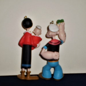 Może przedstawiać: Zestaw trzech świątecznych ozd&oacute;b przedstawiających Popeye'a, Oliwię i ich dziecko. Popeye jest ubrany w sw&oacute;j charakterystyczny str&oacute;j marynarski, Olivia w czerwoną sukienkę, a dziecko w biały kapelusz. Ozdoby są wykonane z plastiku i pomalowane żywymi kolorami.