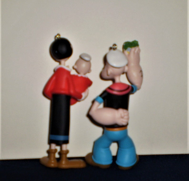 Może przedstawiać: Zestaw trzech świątecznych ozd&oacute;b przedstawiających Popeye'a, Oliwię i ich dziecko. Popeye jest ubrany w sw&oacute;j charakterystyczny str&oacute;j marynarski, Olivia w czerwoną sukienkę, a dziecko w biały kapelusz. Ozdoby są wykonane z plastiku i pomalowane żywymi kolorami.