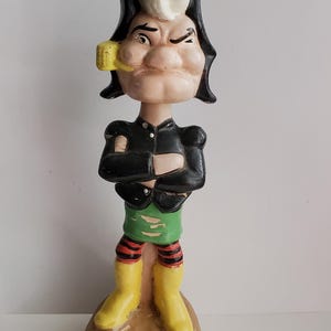 May include: A ceramic figurine of a cartoon character with a black hat, yellow boots, and a green and red striped skirt. The figurine is standing on a base with the text "Lil Abner Dogpatch Enterprises Inc. USA" on it.