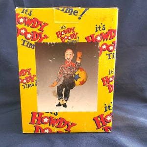 May include: A yellow cardboard box with a cartoon character on the front. The character is a puppet with a plaid shirt, blue jeans, and a cowboy hat. The character is sitting on a yellow ball with blue stars. The box has the words "It's Howdy Doody Time!" printed on it.