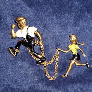 May include: Gold-toned metal brooch featuring two figures, a man and a woman, running. The man is wearing a white shirt, black pants, and black shoes. The woman is wearing a yellow shirt, black skirt, and black shoes. They are connected by a gold chain.