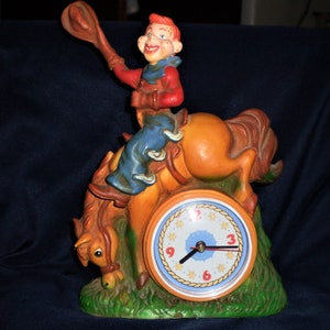 May include: A vintage ceramic clock featuring a cowboy riding a bucking horse. The cowboy is wearing a blue shirt, brown pants, and a red vest. The clock face is blue with white stars and gold numbers.
