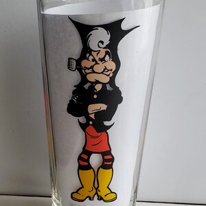 May include: A clear glass pint glass featuring a cartoon character with a black cape, black outfit, red shorts, and yellow boots. The character is smoking a pipe. The word "MAMMY" is printed at the bottom of the glass.