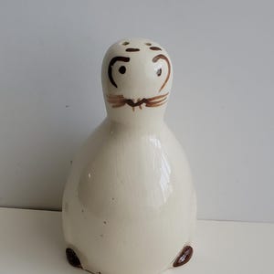 May include: Cream-colored ceramic salt shaker with a seal-like design. The shaker features brown painted details for the eyes, whiskers, and flipper-like feet. The top has three holes for dispensing salt. A whimsical kitchen decor item.