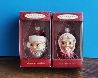Peut inclure: Deux décorations de Noël dans des boîtes. L'une est le Père Noël avec une barbe et un chapeau blancs. L'autre est la Mère Noël avec un bonnet blanc et un nœud rouge. Les boîtes sont rouges avec le texte "Keepsake Ornament" et "Décoration des Fêtes".