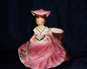 May include: A vintage ceramic figurine of a girl wearing a pink hat and dress, sitting on a tree stump and holding a book. The figurine is a planter with a brown tree stump base.