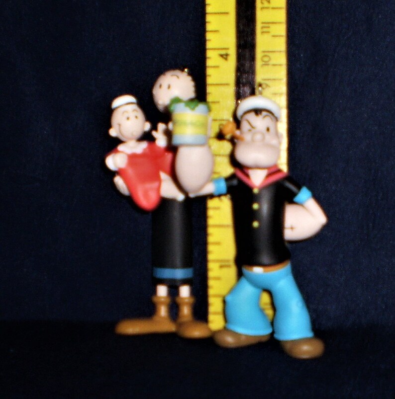 Może przedstawiać: Zestaw trzech figurek Popeye'a. Popeye trzyma puszkę szpinaku i dziecko Popeye'a. Dziecko ma na sobie czapkę marynarską i czerwoną koszulę. Popeye ma na sobie niebieską marynarską koszulę i niebieskie spodnie. Figurki są wykonane z plastiku i mają około 10 cm wysokości.