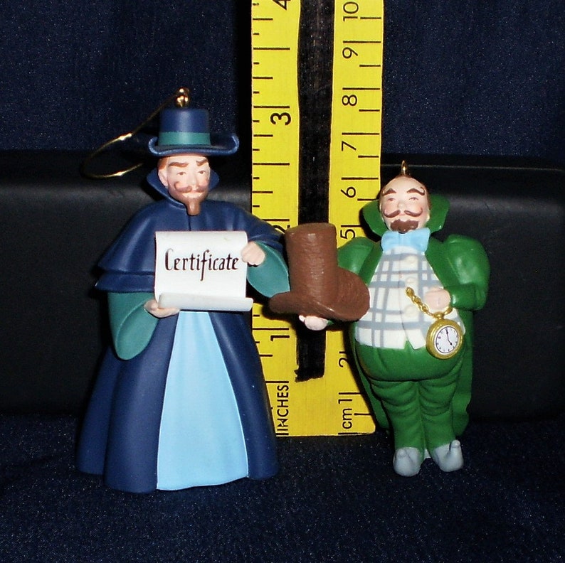Peut inclure: Deux d&eacute;corations de No&euml;l repr&eacute;sentant des personnages du film 'L'&Eacute;trange No&euml;l de monsieur Jack'. La d&eacute;coration de gauche est une figure v&ecirc;tue d'une robe bleue et verte tenant un parchemin qui dit 'Certificat'. La d&eacute;coration de droite est une figure v&ecirc;tue d'une robe verte avec une chemise blanche et une montre de poche.