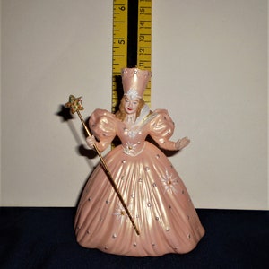 Peut inclure: Une figurine en porcelaine rose de la M&eacute;chante Sorci&egrave;re de l'Ouest du Magicien d'Oz. Elle porte une robe rose avec des &eacute;toiles argent&eacute;es et une couronne blanche. Elle tient une baguette dor&eacute;e surmont&eacute;e d'une &eacute;toile verte.