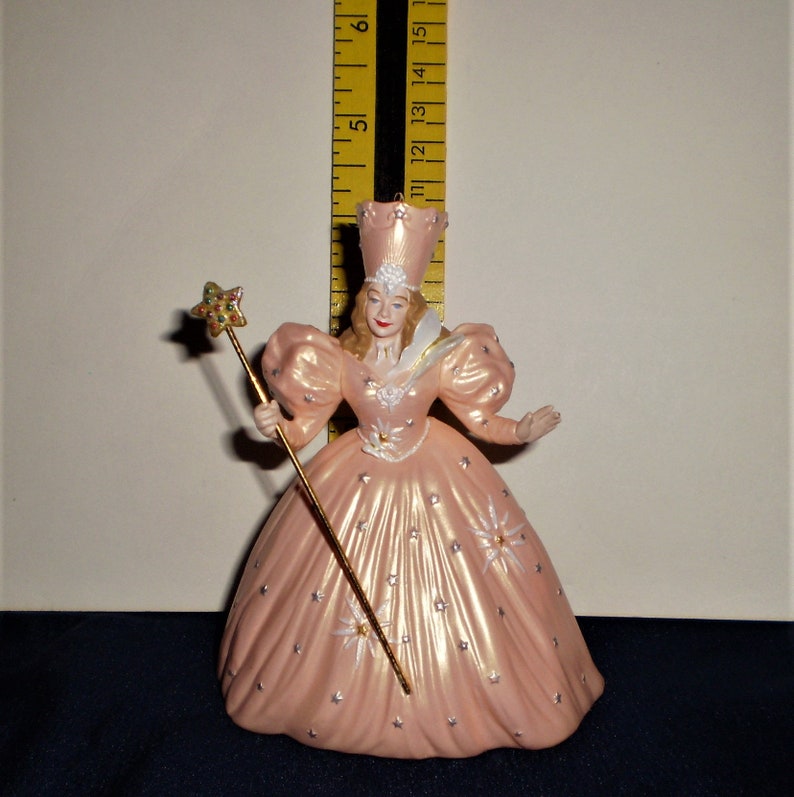 Peut inclure: Une figurine en porcelaine rose de la M&eacute;chante Sorci&egrave;re de l'Ouest du Magicien d'Oz. Elle porte une robe rose avec des &eacute;toiles argent&eacute;es et une couronne blanche. Elle tient une baguette dor&eacute;e surmont&eacute;e d'une &eacute;toile verte.