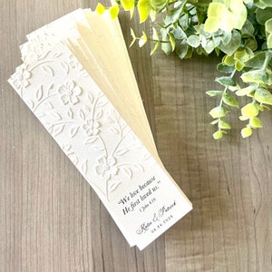 May include: White bookmark with a floral design and the text "We love because He first loved us." 1 John 4:19. Below the text is the couple's names "Katie & Patrick" and the date "08.24.2024".