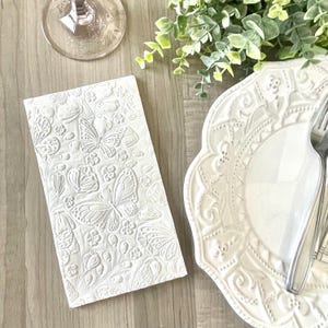May include: White embossed paper napkin with a butterfly and floral design. The napkin is on a wooden table next to a white plate with a decorative edge.