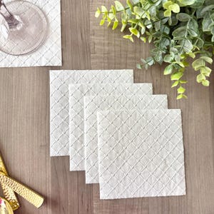 May include: A set of four square white paper napkins featuring an embossed diamond grid pattern with small hearts at each intersection. The napkins are arranged in a slightly overlapping stack, displaying a consistent texture and white color throughout.