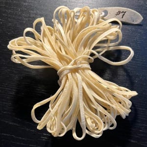 May include: A bundle of cream-coloured, thin, flat leather strips tied in the centre with a matching cord. A small, off-white, pointed paper tag is attached to the top, featuring the handwritten text #7 in black ink on its surface.