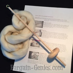 May include: A light-colored wooden drop spindle with a metal hook at the top and a round whorl, accompanied by a coiled bundle of off-white wool roving and a printed instruction sheet titled Bottom Whorl Drop Spindle Instructions.