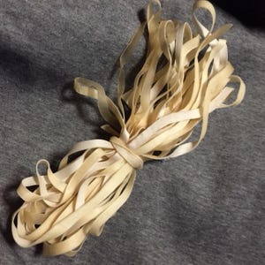 May include: A bundle of cream-colored, thin ribbons tied together. The ribbons are arranged in a loose, flowing pattern, with some loops at the ends. The ribbons appear to be made of a soft, possibly fabric-like material.