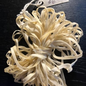 May include: A bundle of thin, off-white elastic bands tied together with a white string. A small, rectangular metal tag is attached to the string, featuring the handwritten text #6 in black ink. The elastic material appears flexible and coiled.