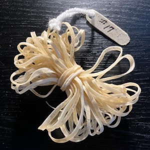 May include: A bundle of light beige, thin, ribbon-like material tied together with a matching band. A small, rectangular tag with the number "#17" is attached with a white string. The items are on a dark surface.