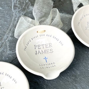 May include: White ceramic ornament with a cross and the text "The Lord bless you and keep you Peter James Baptized 08.07.22"