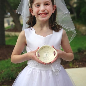 May include: A young girl in a white dress and veil holds a white ceramic dish with the words "Gianna Therese" and "First Holy Communion" written on it. The dish has a small cross design.