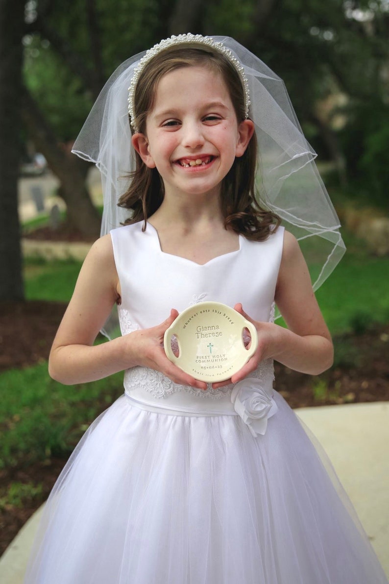 May include: A young girl in a white dress and veil holds a white ceramic dish with the words "Gianna Therese" and "First Holy Communion" written on it. The dish has a small cross design.