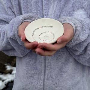 A woman in a purple jacket is holding a white ceramic bowl featuring the nine Fruits of the Holy Spirit