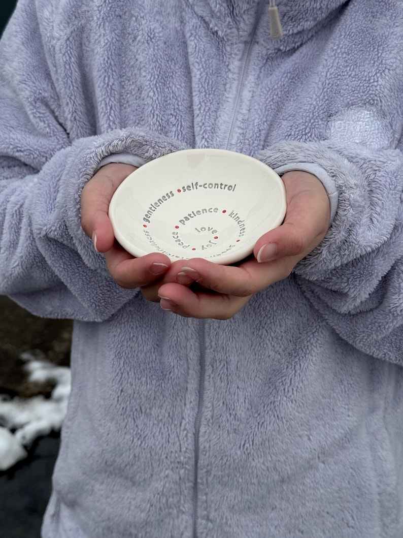 A woman in a purple jacket is holding a white ceramic bowl featuring the nine Fruits of the Holy Spirit