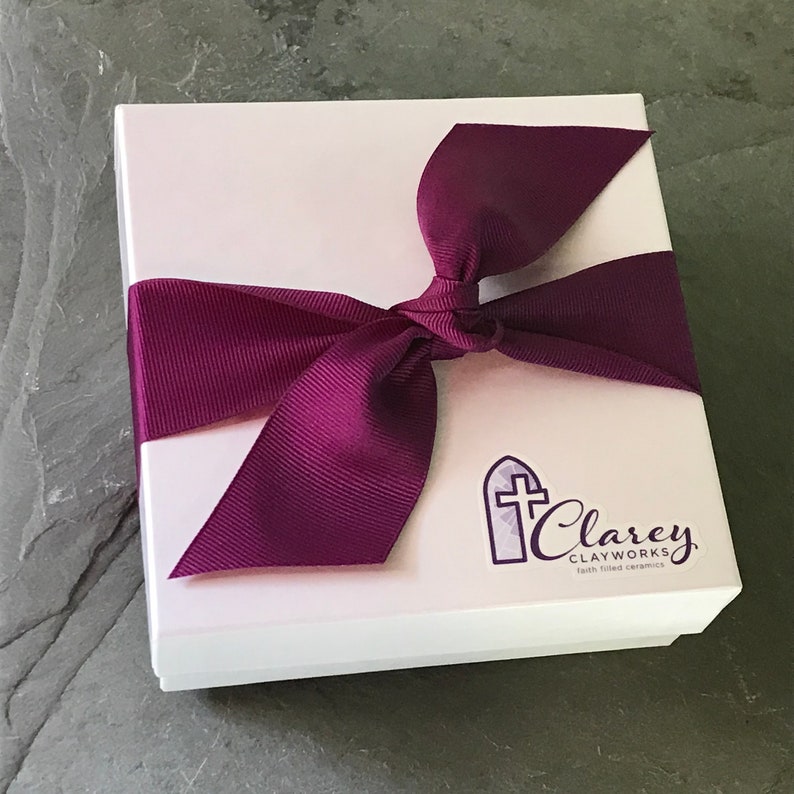 Peut inclure: Une bo&icirc;te cadeau blanche avec un ruban violet nou&eacute; en un n&oelig;ud. La bo&icirc;te porte un logo qui dit "Clarey Clayworks" avec une croix et un motif de fen&ecirc;tre d'&eacute;glise. Le texte sous le logo dit "c&eacute;ramiques remplies de foi".