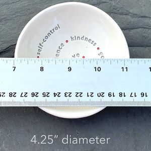 A white ceramic bowl featuring the nine Fruits of the Holy Spirit is shown from a top view with a ruler across showing 4.25" diameter.