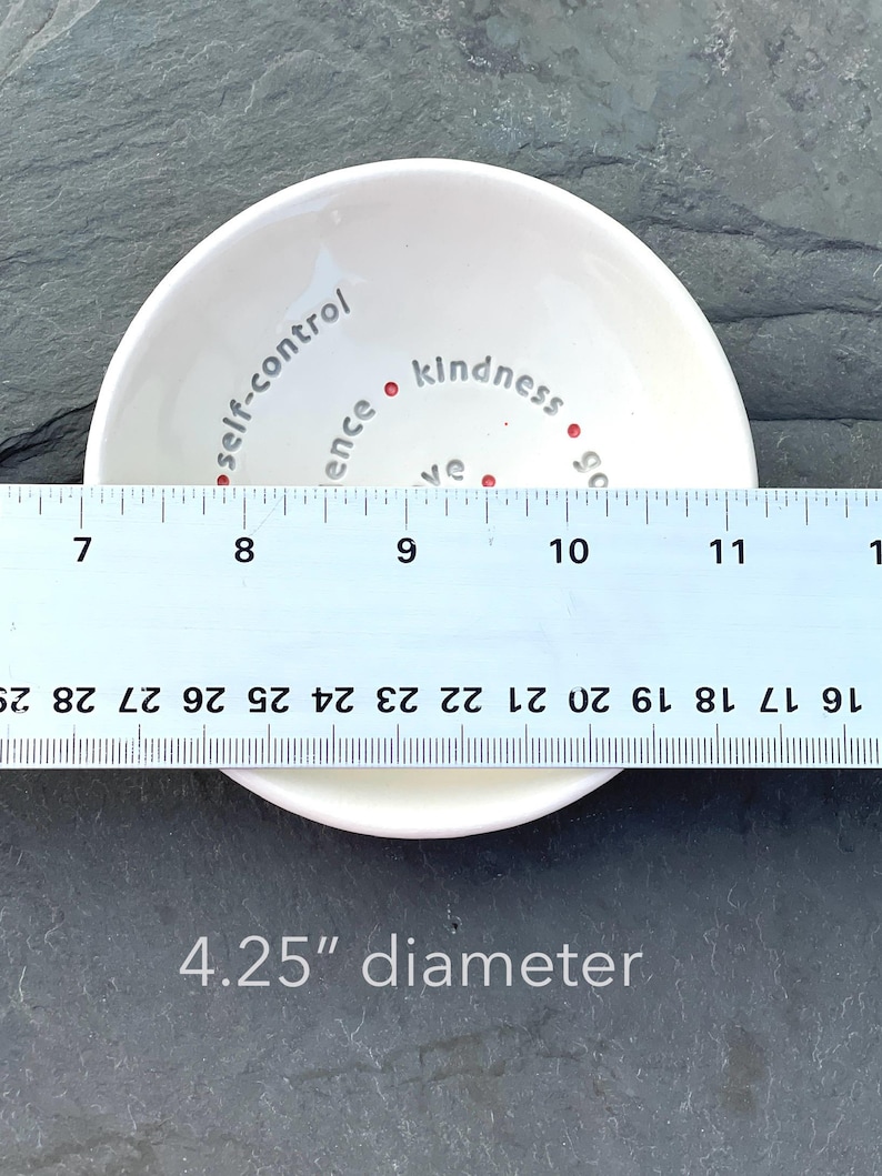 A white ceramic bowl featuring the nine Fruits of the Holy Spirit is shown from a top view with a ruler across showing 4.25" diameter.